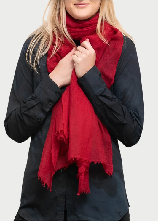 A person with long, blonde hair, wearing a black shirt and a luxurious Whisperweight Cashmere Scarf by Meg Cohen stands against a plain white background. They hold the scarf close to their neck with both hands and gaze at the camera with a neutral expression.