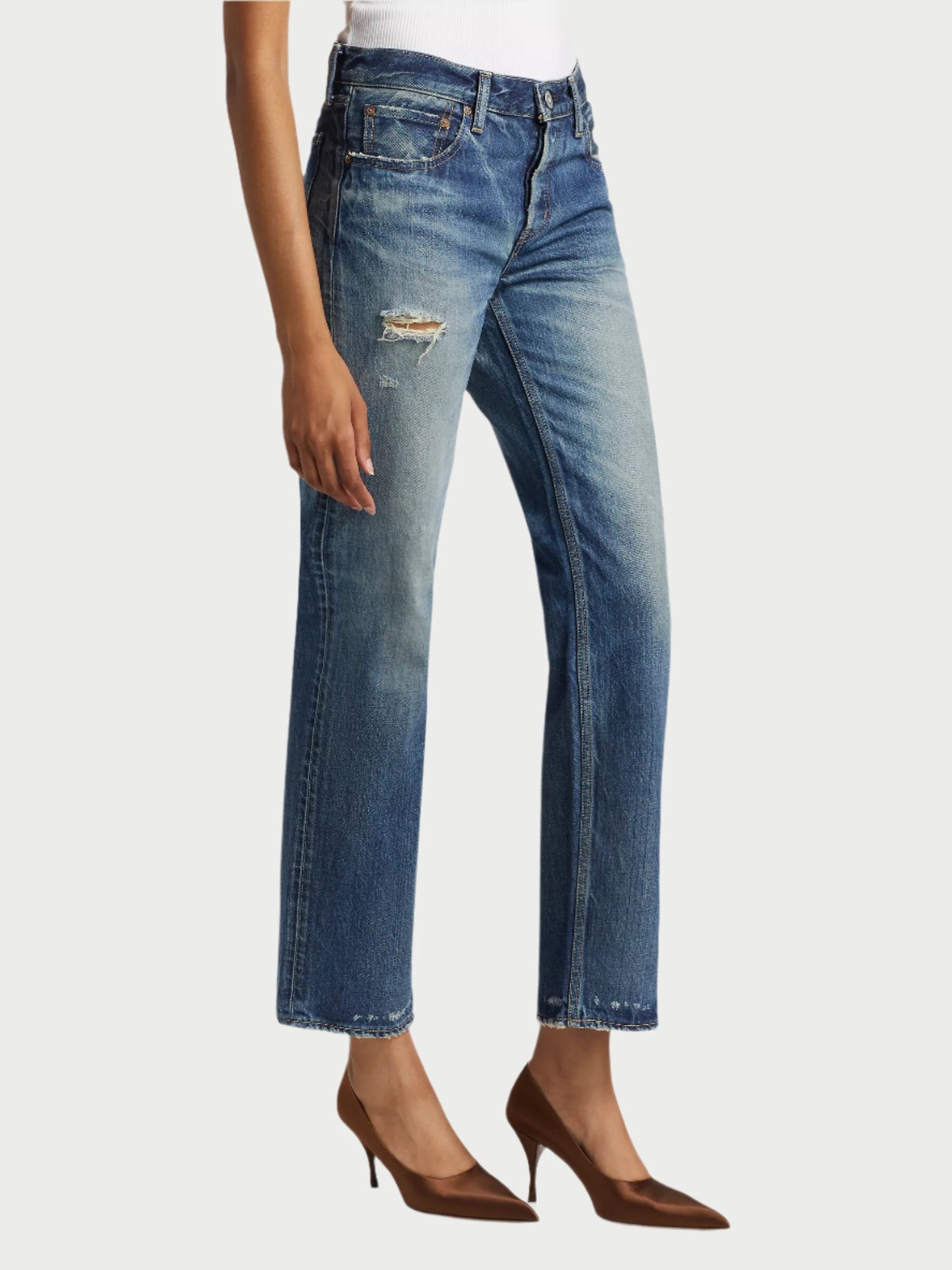 A person sports the stylish Flemington Straight Low Rise Jeans by Moussy paired with a white top and brown high heels. These jeans feature a chic rip on the upper thigh and maintain a straight-leg fit, adding an effortlessly sophisticated vibe to the ensemble.