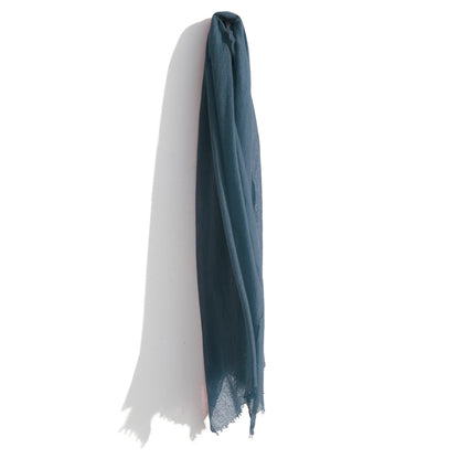 A lightweight, teal-colored Whisperweight Cashmere Scarf by Meg Cohen hangs against a white wall. The scarf is positioned vertically and its bottom edges are frayed. Shadows are cast on the wall behind it, adding depth to the image. The natural fibers of the scarf give it an elegant and soft appearance.