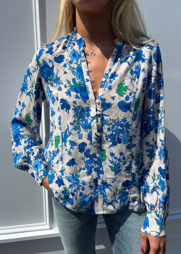 A person stands against a light-colored background wearing the Twina Crepe Blouse by Zadig & Voltaire, which showcases blue and green floral prints on white crepe viscose fabric. The blouse, featuring buttons down the front, is machine washable. With one hand casually in their jeans pocket, the person's face is not visible.