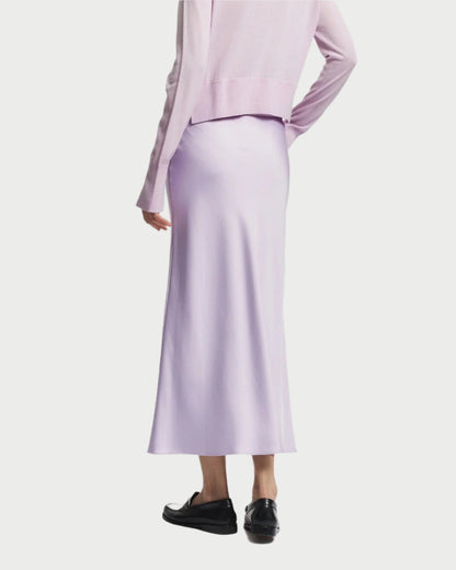 Rear view of a person in a light purple, long-sleeve top with a matching Saint Art NY Charmeuse Maxi Skirt. They stand with one hand on their hip, highlighting the sleek bias cut while wearing black loafers.