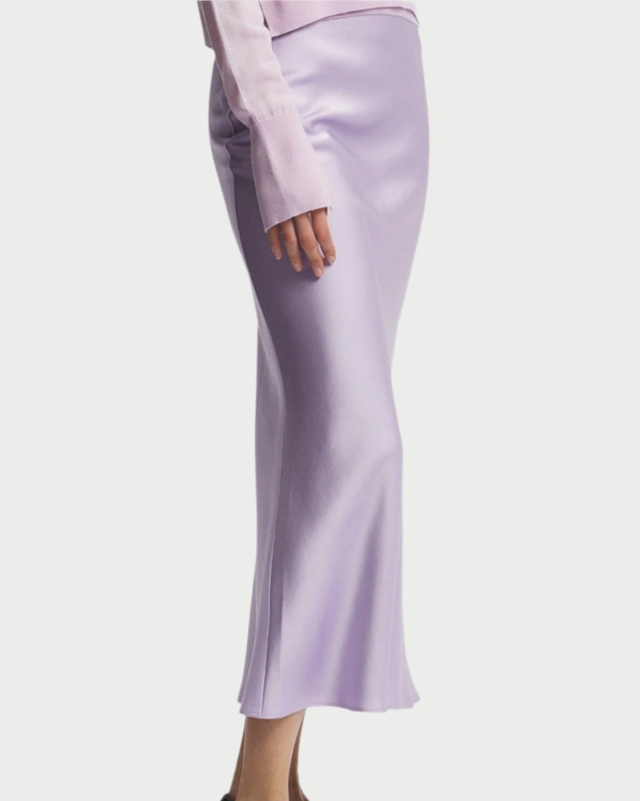 A person is wearing a pale pink long-sleeved top paired with a Saint Art NY Charmeuse Maxi Skirt in lavender, delightfully draped on the bias. Black loafers with white accents complete the stylish ensemble against a plain white background.