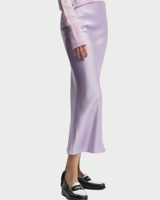 A person is wearing a pale pink long-sleeved top paired with a Saint Art NY Charmeuse Maxi Skirt in lavender, delightfully draped on the bias. Black loafers with white accents complete the stylish ensemble against a plain white background.