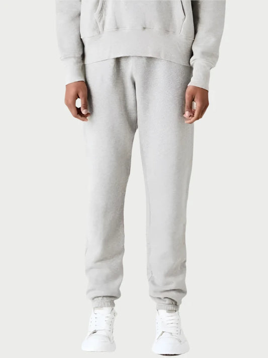 A person is standing against a plain white background, dressed in an elevated and comfortable LES TIEN ensemble. They sport a light gray Zendaya Hoodie with front pocket, paired with Dylan Classic Sweatpants and white sneakers. The outfit radiates a relaxed yet stylish vibe.