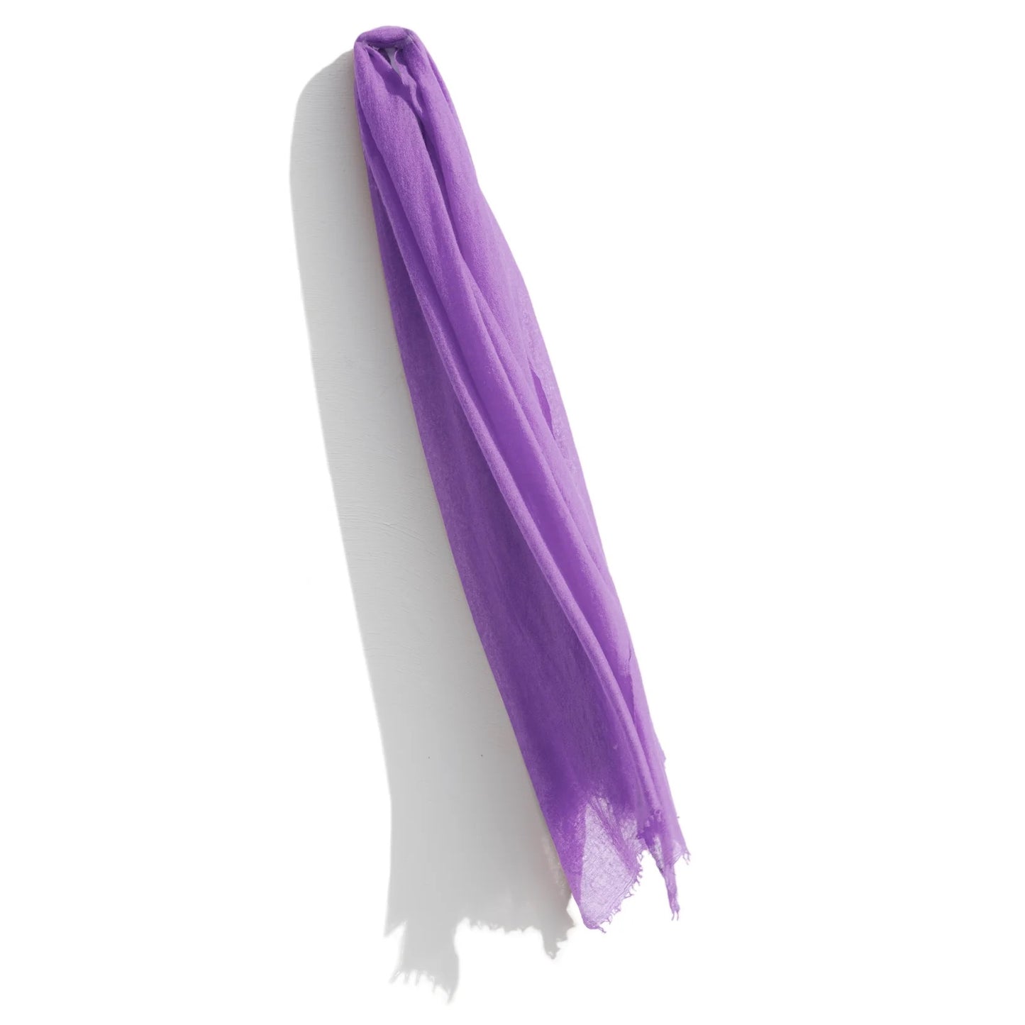 A sheer, lightweight purple Whisperweight Cashmere Scarf by Meg Cohen is draped elegantly against a white background, with its ends slightly frayed. The scarf's natural fibers create subtle folds and shadows, adding texture to the simple yet striking composition.