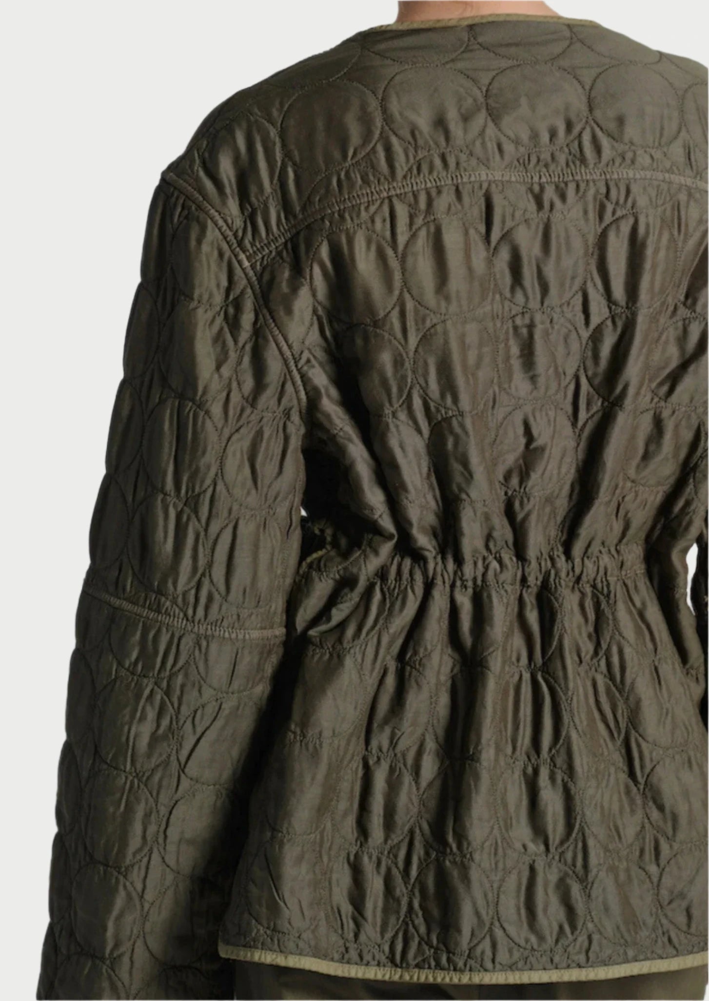 A person is viewed from behind wearing a Quilted Jacket by Saint Art NY in olive green. The jacket displays circular stitching patterns and includes a cinched waist for a customizable fit, blending style and comfort seamlessly. The background is plain light gray.