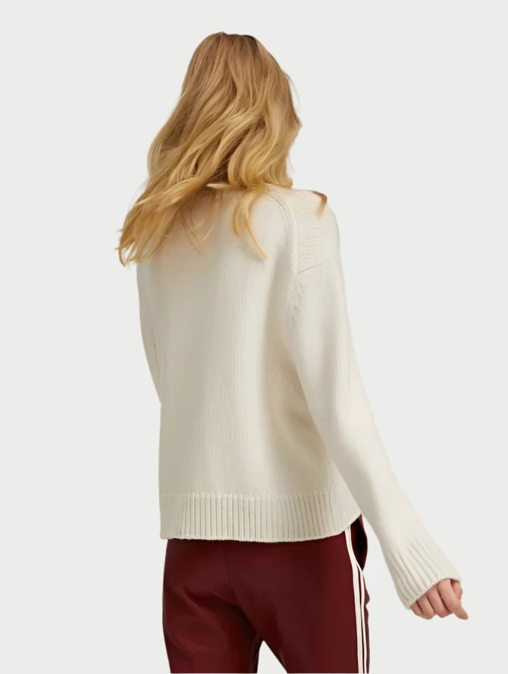 A person with long blonde hair is shown from the back, wrapped in the cozy Ivory Heavy Crewneck Sweater by SPRWMN and wearing maroon pants with white stripes on the side. The background is a plain light gray.