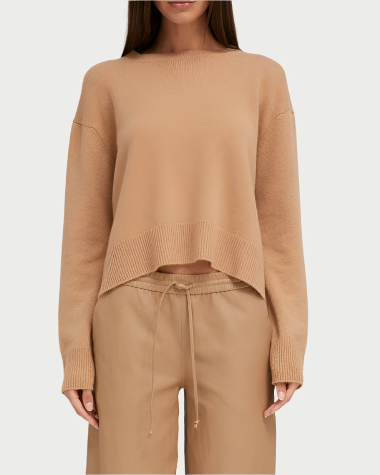 A person is wearing SPRWMN's Boxy Praline Cashmere Sweater with drawstring pants. The light brown sweater has a relaxed fit and high-low hem, with the front slightly tucked in. An off-white background complements the cozy look.