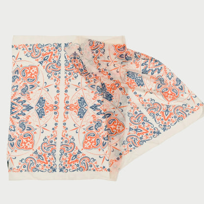 A large rectangular cotton voile scarf from Maison Lecomte Flament features an intricate paisley pattern in orange, blue, and white. The design boasts symmetrical elements with detailed motifs, and the partially folded scarf showcases both front and back sides, making it a captivating creation.