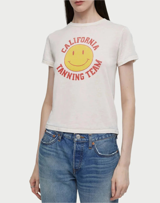 A person with long dark hair wears the RE/DONE "California Tanning Team" T-shirt, featuring a cheerful smiley face. Paired with jeans, they stand against a plain background, effortlessly showcasing casual style.