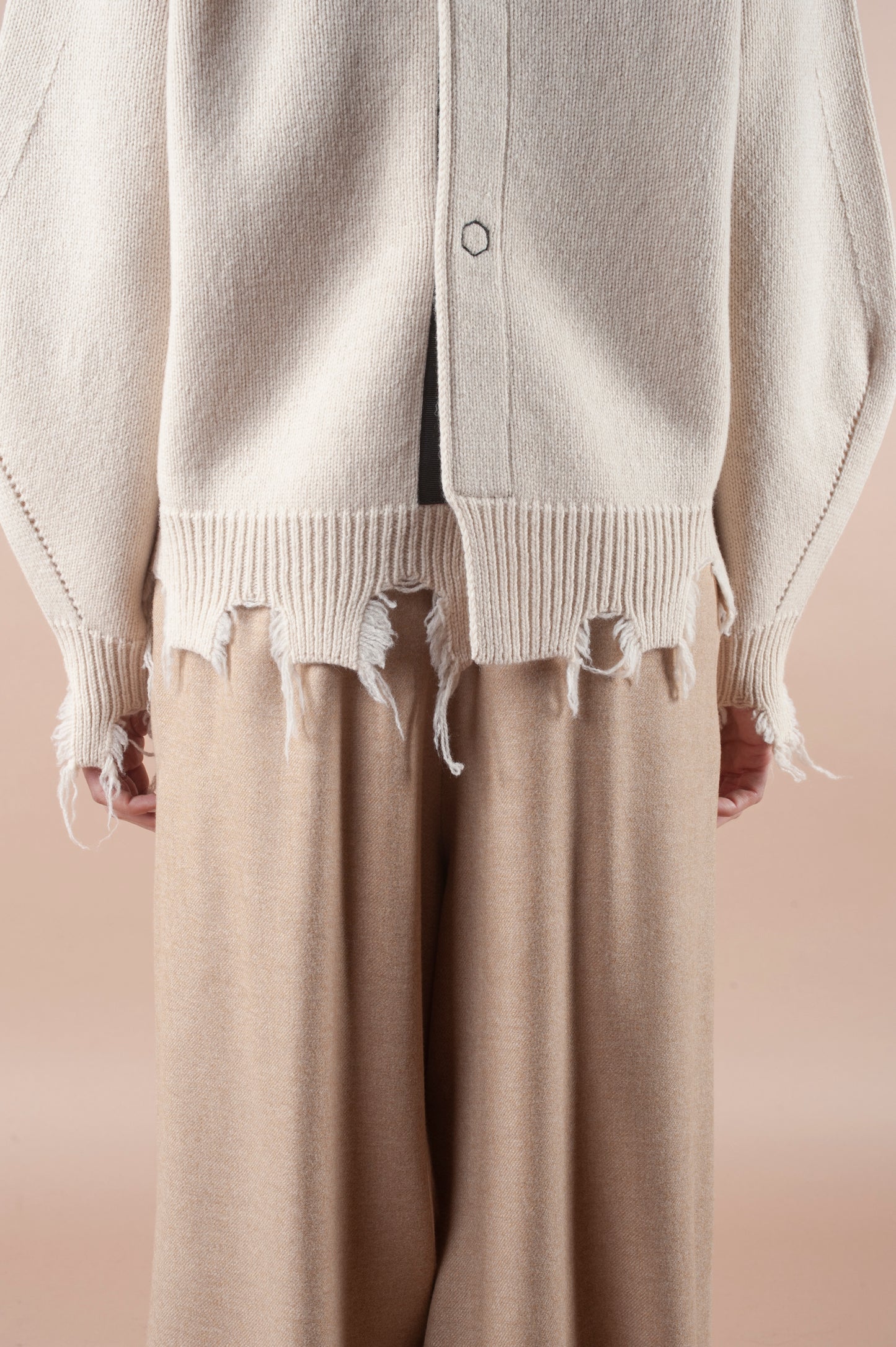 A person wearing the Phisique du Role's Turnable Sweater with Banana Sleeves, featuring a cream hue and unique frayed hem, is paired with loose-fitting beige pants. The sweater boasts a split back and visible stitching detail, all set against a neutral beige background.