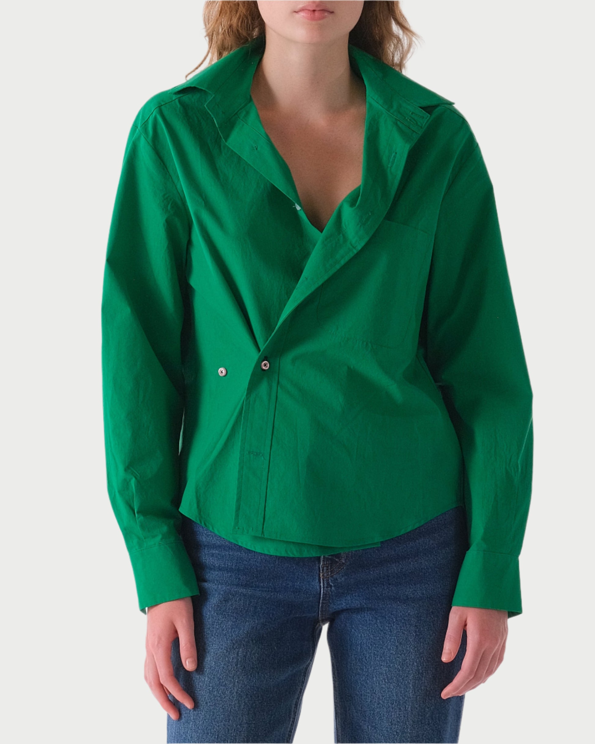 A woman models The Daily Shirt by WEAR CISSA, a vibrant green, menswear-style piece made from luxurious Egyptian cotton. It's partially buttoned over blue jeans, complementing her understated pose against a plain white backdrop, with hands casually by her sides and face unseen.