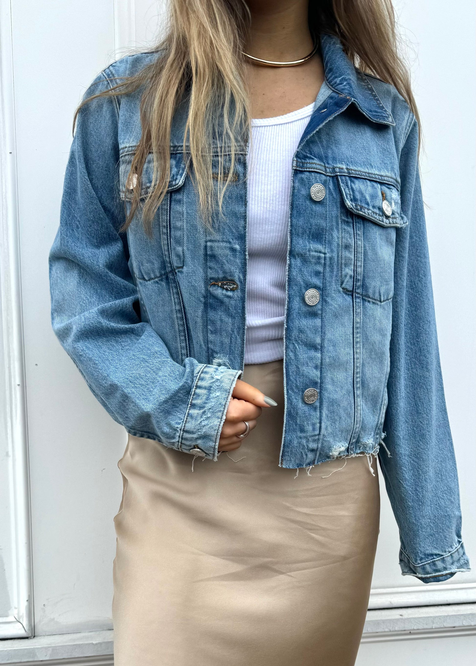 A person with long blonde hair wears a stylish Frame Vintage Denim Distressed Jacket over a white ribbed tank top and a beige satin skirt. They have a neutral-colored manicure and accessorize with a simple silver necklace, set against the backdrop of a plain white wall.