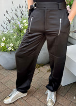 Load image into Gallery viewer, A person is wearing shiny black Charmeuse Pants by Saint Art NY with silver zippers on the front pockets, a black top, and white sneakers with gold accents. They are standing on a paved area near large gray planters filled with lavender and other flowers.