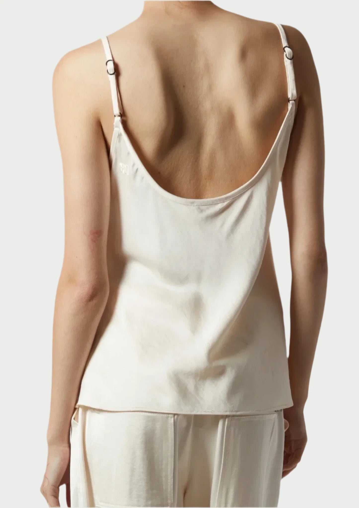 A person is wearing a cream-colored, silky-smooth Saint Art NY Charmeuse Cami with thin straps and a low back. The person is facing away from the camera, highlighting the modern femininity of the top and matching cream-colored pants against a plain, neutral background.