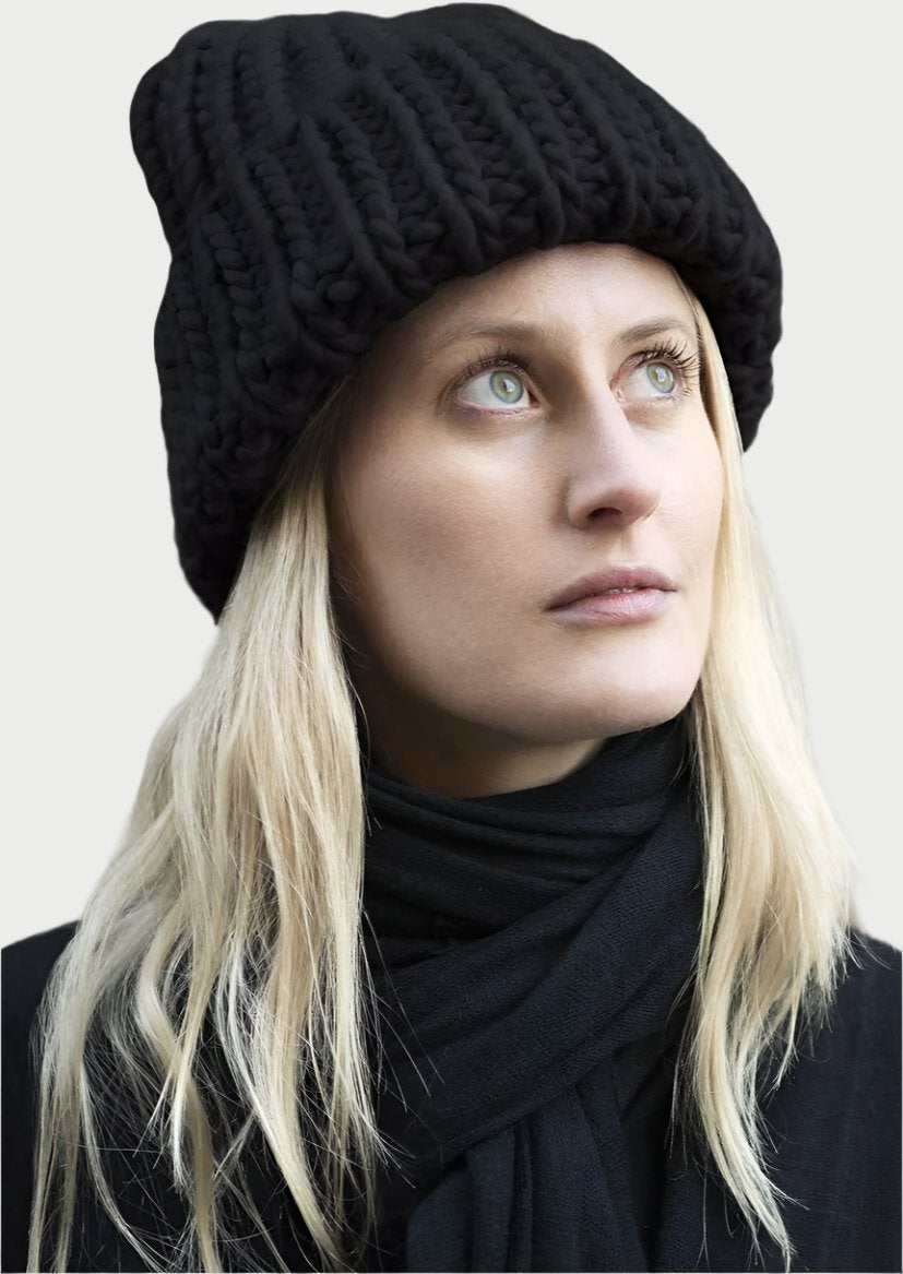 A person with long blond hair is wearing a thick black knitted hat made from a Soft Wool Beanie by Meg Cohen and a matching black scarf. They are looking slightly upwards against a neutral background.