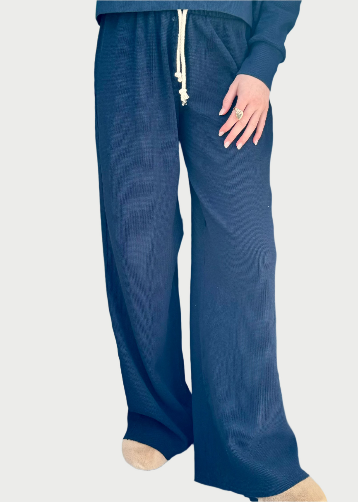 A person standing on a wooden walkway wearing dark blue Kylie Waffle Pant by perfectwhitetee wide-leg loungewear with a drawstring waist and tan shoes. The person's right hand is visible, showing a ring on their finger, while the rest of their upper body is mostly out of the frame.