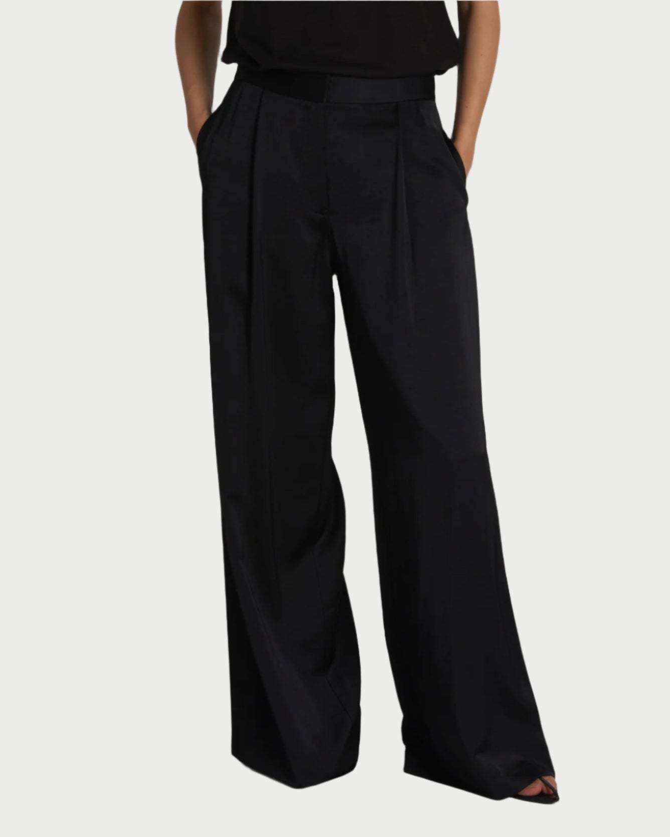 A person wears Saint Art NY's Nave Black Wide Leg Pants, characterized by their loose fit, high waist, and elegant charmeuse fabric. Their hands are in pockets. The dark shirt they wear blends with a plain background, though it is not fully visible.