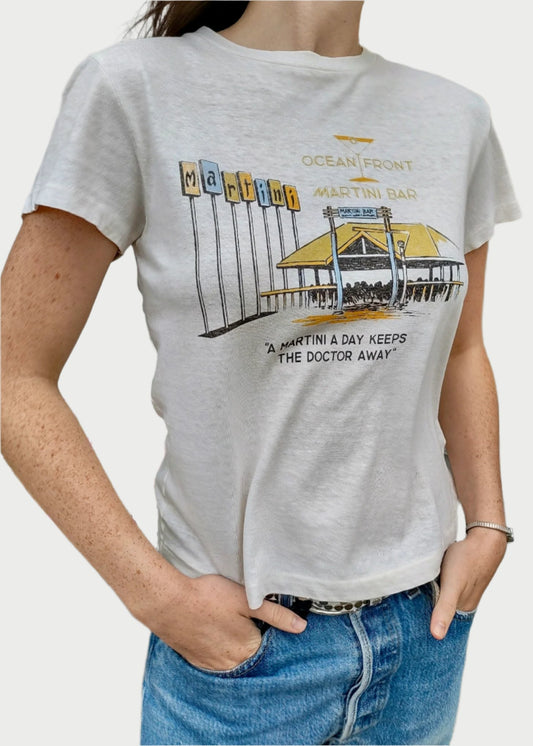 A person wearing a RE/DONE Martini Bar Classic Tee made of premium cotton fabric, showcasing a retro-style building, palm trees, and a sign that says "Ocean Front Martini Bar." The text reads, "A martini a day keeps the doctor away." The person has their hand in the pocket of blue jeans.