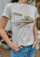 Load image into Gallery viewer, A person wearing a RE/DONE Martini Bar Classic Tee made of premium cotton fabric, showcasing a retro-style building, palm trees, and a sign that says &quot;Ocean Front Martini Bar.&quot; The text reads, &quot;A martini a day keeps the doctor away.&quot; The person has their hand in the pocket of blue jeans.