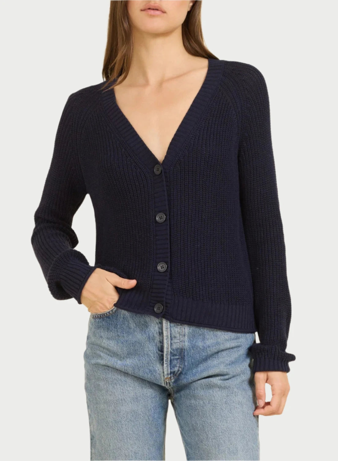 A woman with long, brown hair is donning a deep V-neck, button-up Raleigh Cardigan by One Grey Day, crafted from soft cotton fabric with ribbed detailing in a rich navy hue. She has one hand tucked into the pocket of her light blue jeans against a plain beige background.