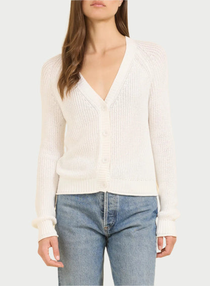 A woman with long brown hair stands against a beige background, wearing an essential piece from One Grey Day: the Raleigh Cardigan, a fitted, buttoned white garment with a deep V-neck made from soft cotton fabric. She pairs it with light blue jeans and has a neutral expression as her hands rest by her sides.