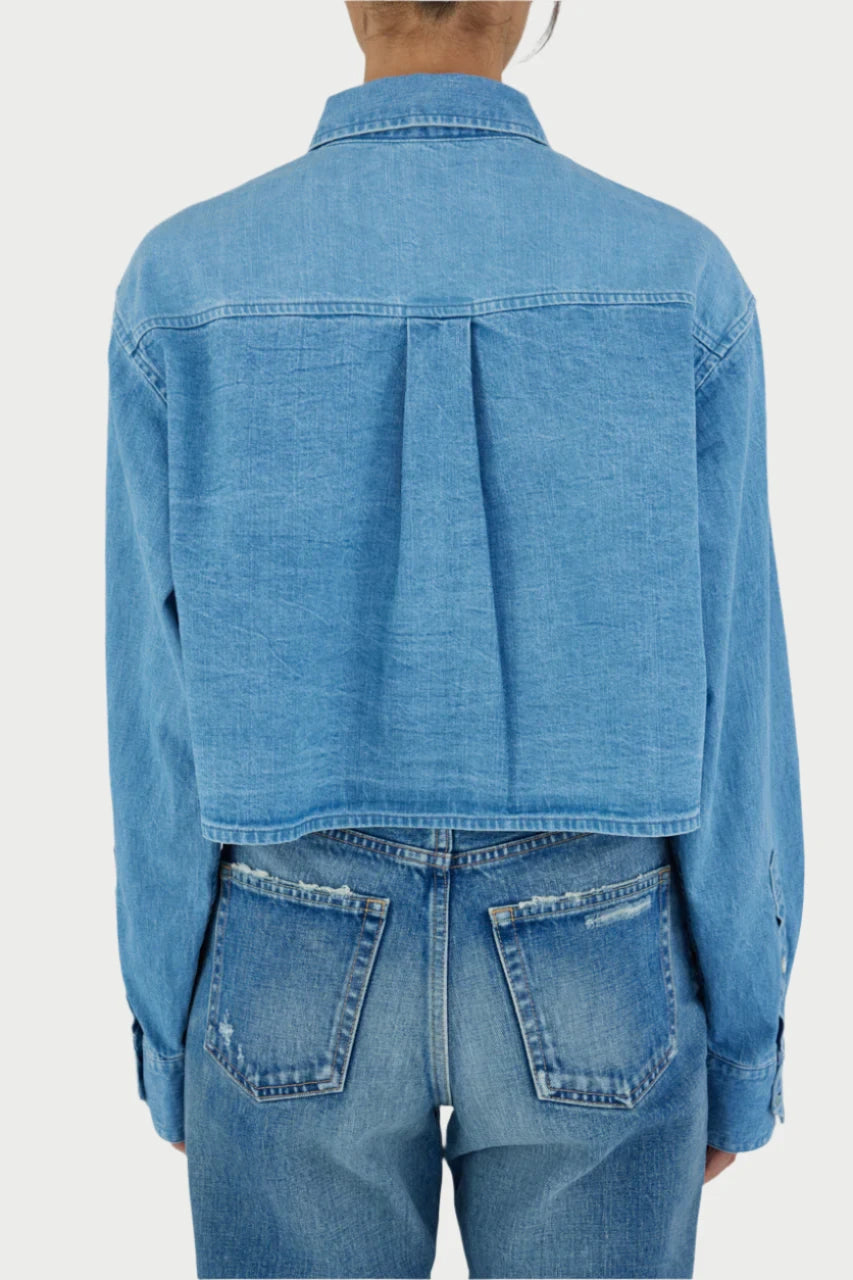 A person is shown from behind wearing a light blue, long-sleeved denim Lenfred Cropped Blocking Shirt by Moussy and blue jeans. The shirt is slightly cropped with a visible seam down the center of the back and button closure. The jeans have a classic five-pocket design. The person's hair is tied back.