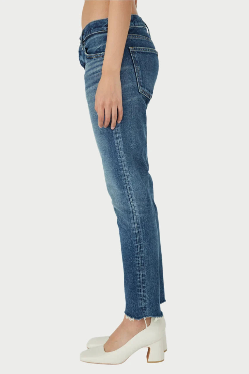 Side view of a person wearing blue Moussy Velden Tapered jeans with a button fly and white heeled shoes. The slightly cropped jeans feature a frayed hem, and the person is standing against a plain white background.