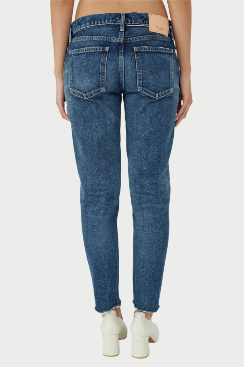 A person is seen from the back wearing Moussy's Ivanhoe Tapered Mid Rise Jeans in blue denim, made from high-quality Japanese fabric for a flattering fit. The jeans feature frayed hems and include two back pockets, paired with white shoes and showcasing a tan label on the waistband.