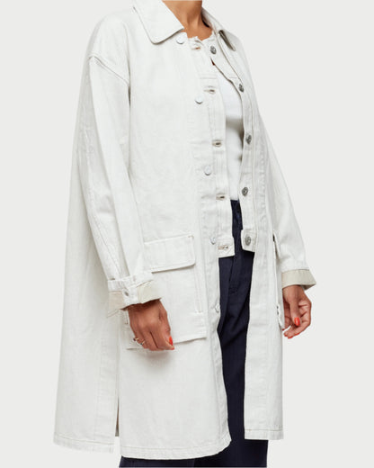 A person is styled in the Hanson Layered Jacket by Moussy, an off-white denim knee-length piece with large pockets, paired with dark pants. The unbuttoned jacket reveals a light shirt underneath, and red nail polish adorns the left hand—ideal for spring transitions.