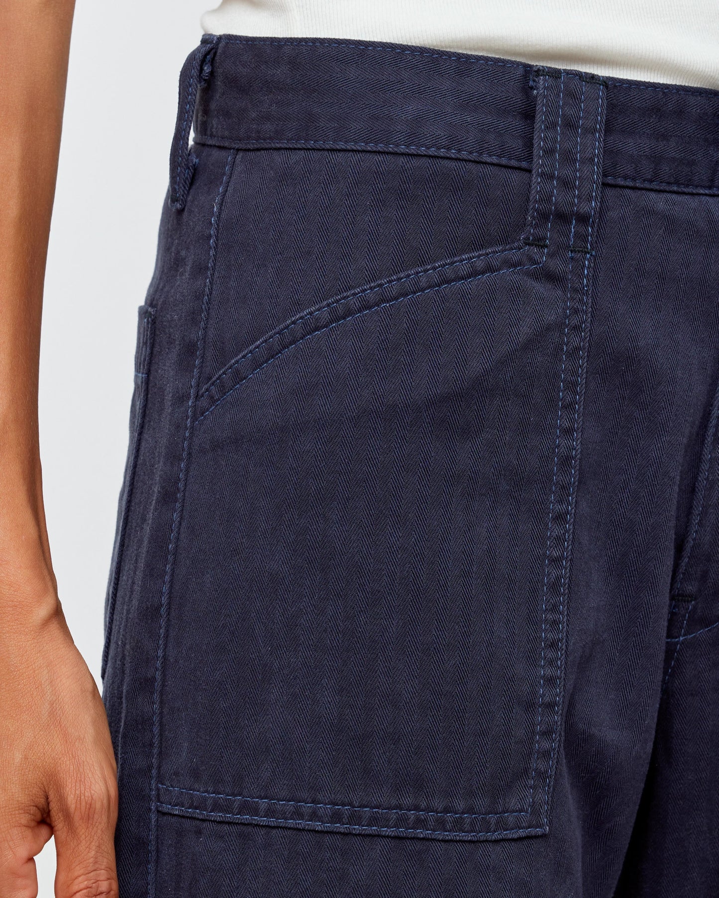 Close-up of an individual in Moussy's Crotona Cargo Pants, a dark blue high-rise alternative to denim featuring visible side pockets and seams. A hand is partially visible near the waist against a plain, light-colored background.