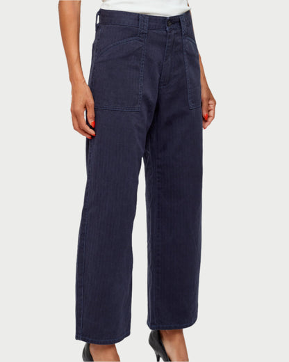 Sporting dark blue Moussy Crotona Cargo Pants, featuring large square front pockets, a white tucked-in shirt complements the high-rise style with visible stitching. Paired with black shoes, the person has one hand casually tucked in a pocket, offering a chic denim look.