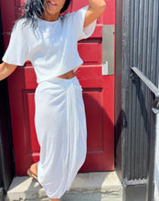 Load image into Gallery viewer, A person wearing a white, midriff-baring two-piece outfit stands in front of a red door with black trim. Sporting dark, wavy hair and one arm raised to their head, they showcase a White Party Sequin Tee from Le Superbe while partially leaning on the door frame and railing.