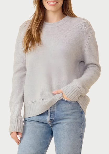 A person with long, brown hair smiles at the camera while wearing the Karyna Cashmere Sweater from One Grey Day and blue jeans. They are standing against a beige background.