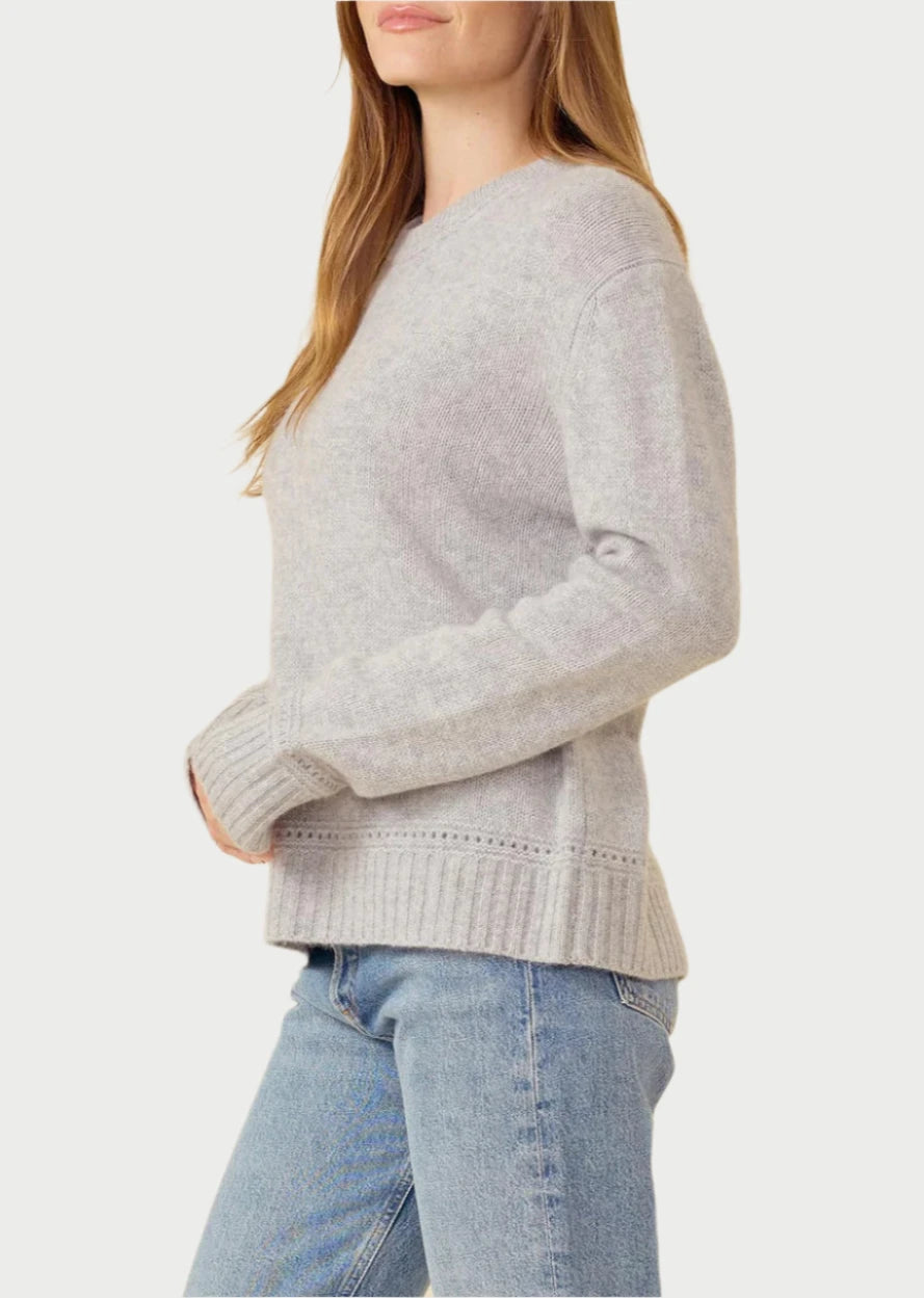 A person with long light brown hair wearing the Karyna Cashmere Sweater by One Grey Day stands in a three-quarter view, facing left. The light gray 100% cashmere sweater showcases unique crochet details on the ribbed cuffs and hem. They pair it with blue jeans and have a neutral expression, set against a plain beige background.