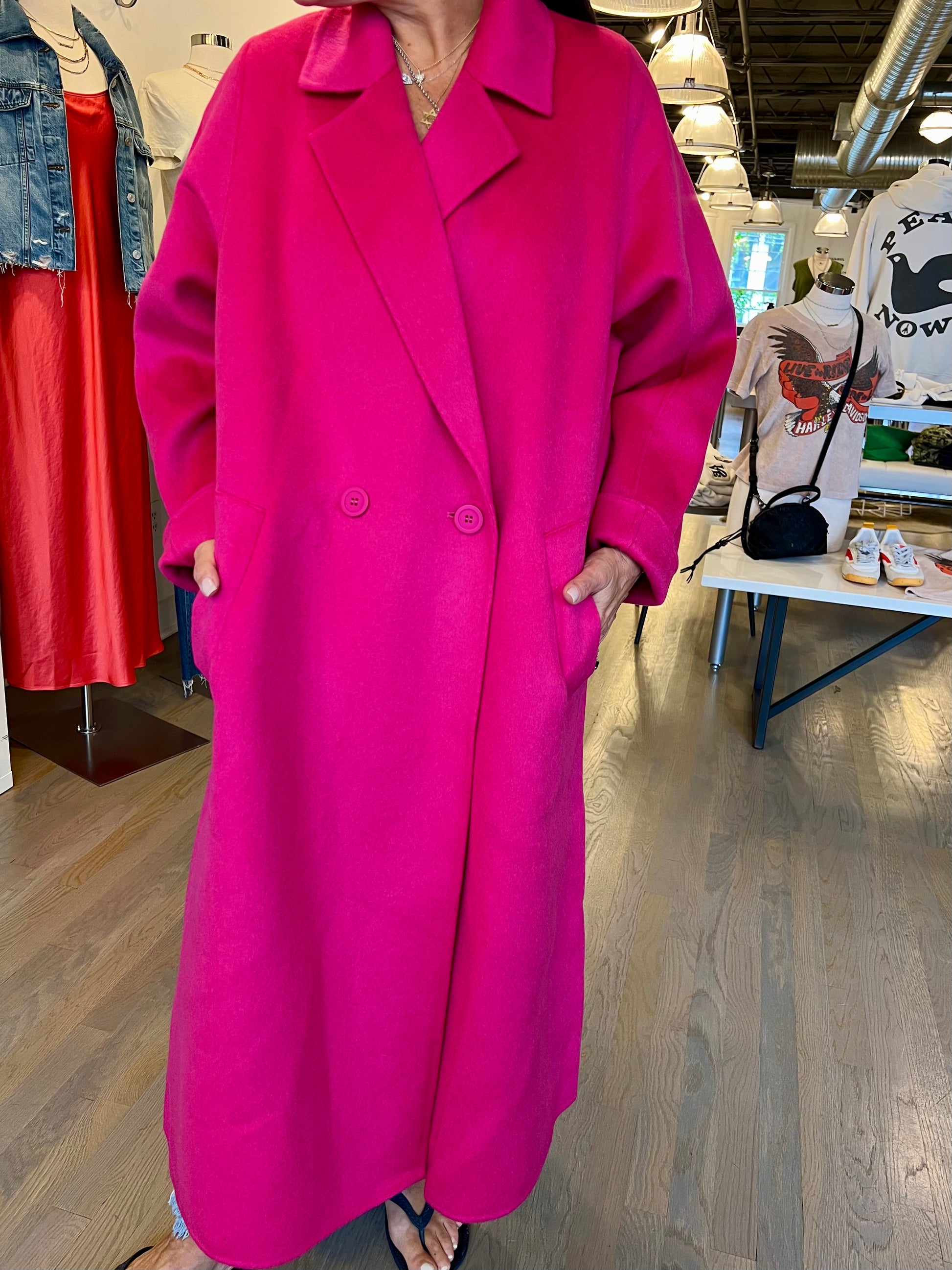 A person wearing a long, bright pink Double Face Coat by Essentiel Antwerp stands indoors. The pure wool coat features large lapels and two visible buttons. In the background, other clothing items are on display, including a red dress, denim jacket, and printed t-shirts.