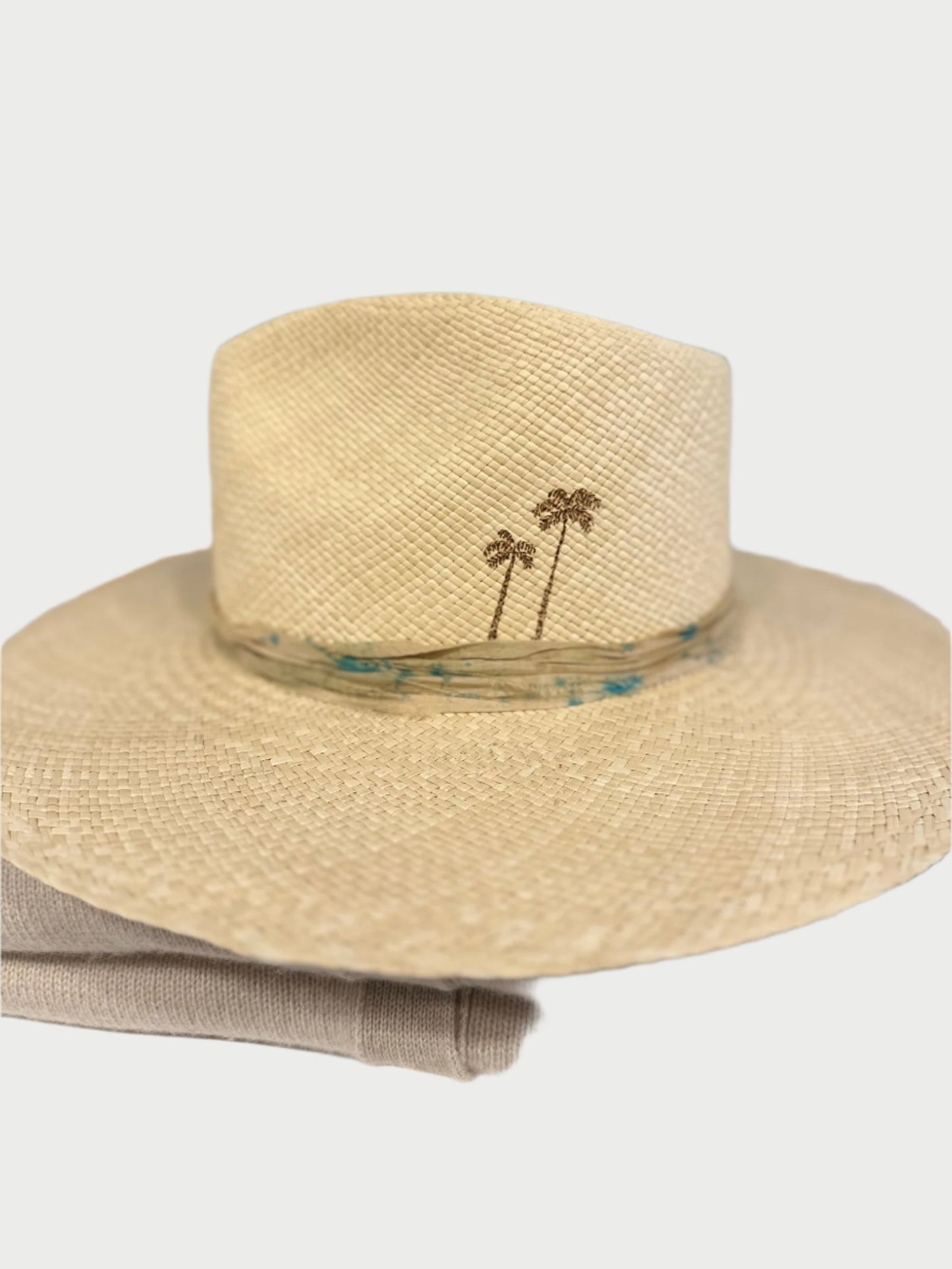 A beige woven straw hat with a wide brim, handmade in the USA by skilled artisans, is displayed on a stack of folded clothes. The Bohemian Glow Straw Hat by Label Jae features a small embroidered design of two palm trees on the front and a light-colored band around the base of the crown. The background is blurred, showing shelves.