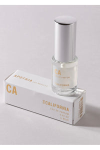 A clear, cylindrical bottle of CA Travel Eau de Parfum (12 ml) is positioned next to its white and gold box, which has "Apothia" printed on it. The bottle has a smooth, shiny silver cap. The light grey surface background highlights the promise of zesty grapefruit and sandalwood notes inside.