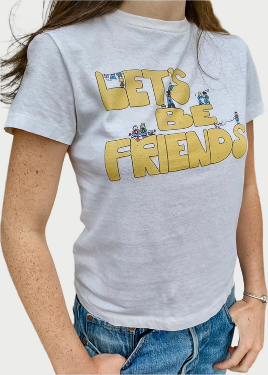 A person wearing the RE/DONE Lets Be Friends Classic Tee, featuring playful yellow text "LET'S BE FRIENDS" adorned with small cartoon characters engaged in various activities on the front. The versatile white t-shirt adds charm to their look as they stand outdoors in front of a potted plant with pink flowers.