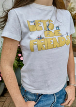 Load image into Gallery viewer, A person wearing the RE/DONE Lets Be Friends Classic Tee, featuring playful yellow text &quot;LET&#39;S BE FRIENDS&quot; adorned with small cartoon characters engaged in various activities on the front. The versatile white t-shirt adds charm to their look as they stand outdoors in front of a potted plant with pink flowers.