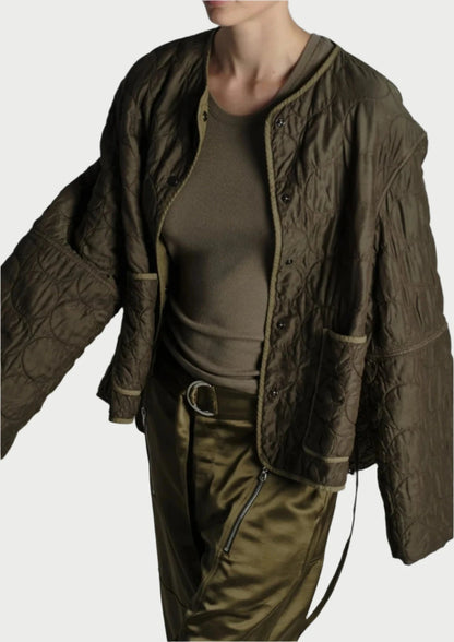 A person wearing a dark green, oversized, quilted jacket from Saint Art NY with wide sleeves, paired with an olive green fitted top and matching olive green pants poses against a plain light background. The outfit exudes a relaxed and stylish look with its military-inspired color scheme and customizable fit.