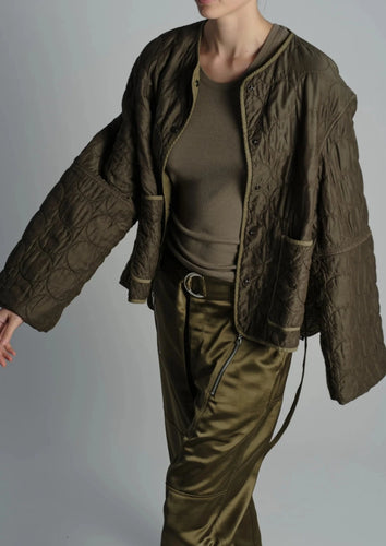 A person wearing a dark green, oversized, quilted jacket from Saint Art NY with wide sleeves, paired with an olive green fitted top and matching olive green pants poses against a plain light background. The outfit exudes a relaxed and stylish look with its military-inspired color scheme and customizable fit.
