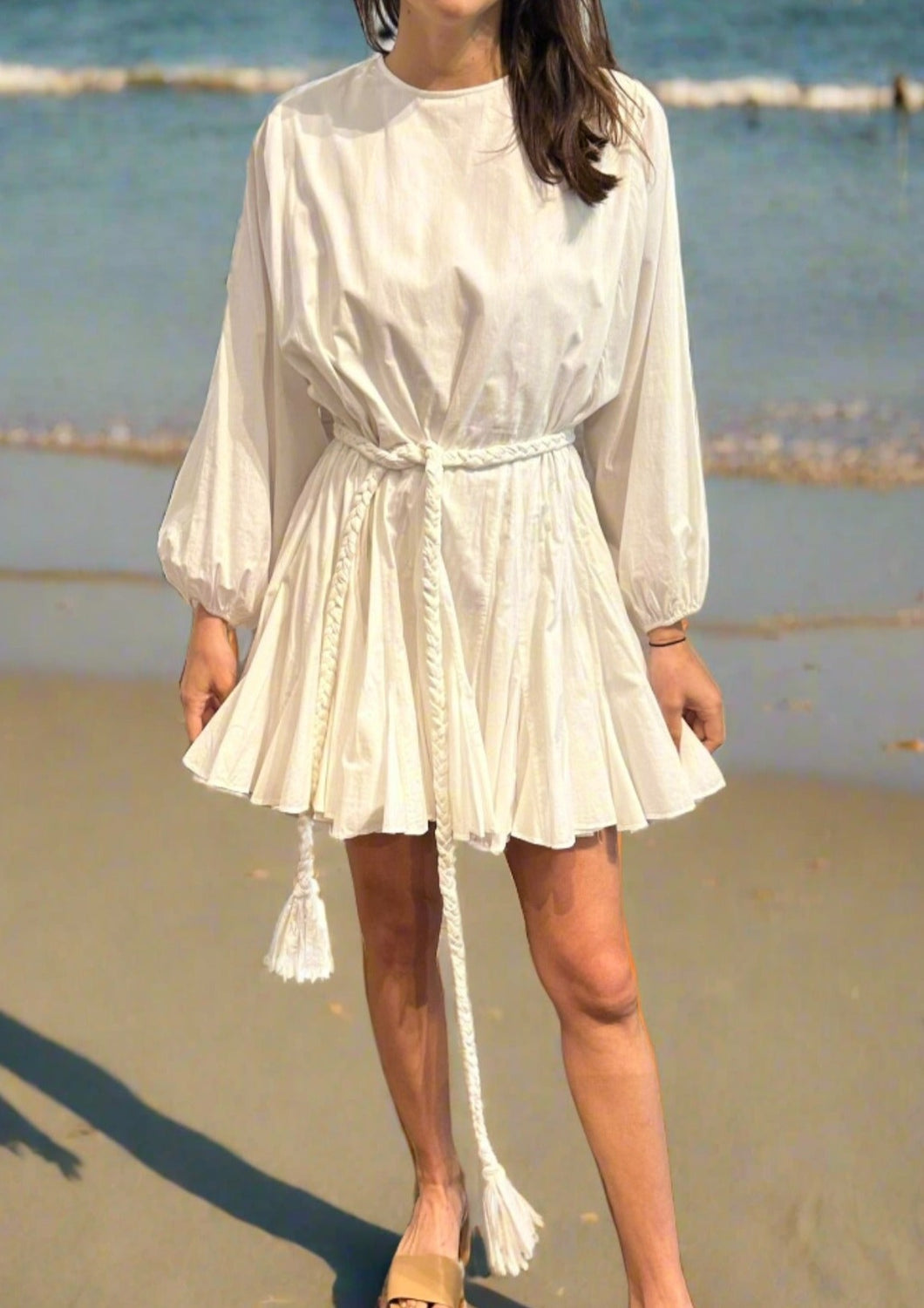 A person stands on a sandy beach near the water, wearing a white, long-sleeved Ella Dress with tassels hanging from the waist. The dress has a pleated skirt and a braided belt, paired with light-colored sandals. Only the lower half of the face is visible.