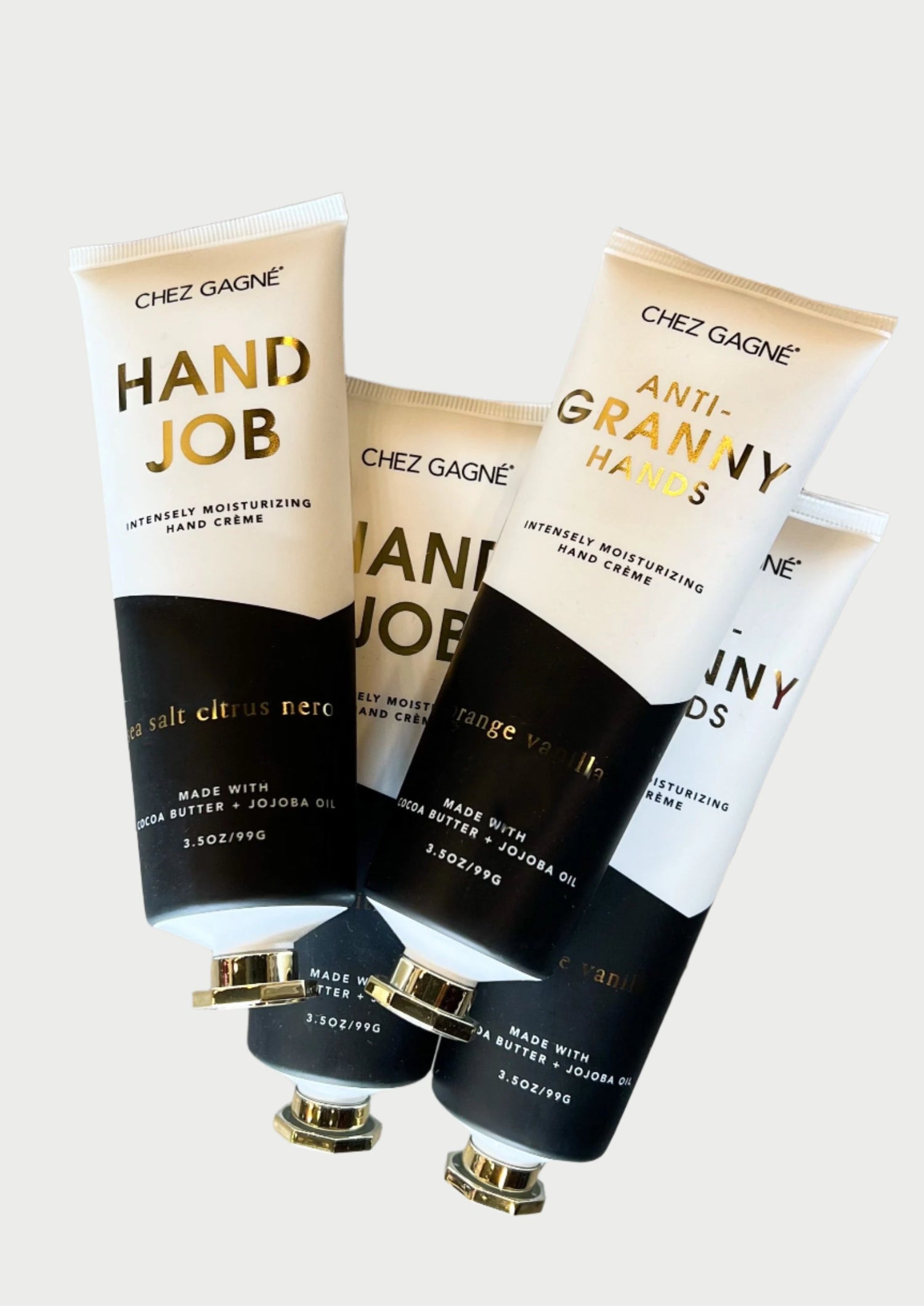 Three tubes of intensely moisturizing Hand Creme on a wooden surface. The tubes display labels with text: "HAND JOB" and "ANTI-GRANNY HANDS," both from the brand Chez Gagné. Infused with cocoa butter and jojoba oil, the scents mentioned are "sea salt citrus neroli" and "orange vanilla.