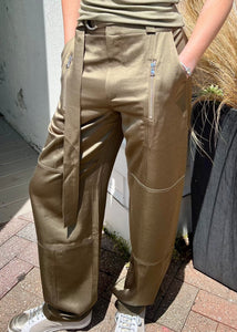 A person is standing with hands in pockets, wearing Saint Art NY's Charmeuse Pant, which are olive green and cargo-inspired with a tied belt and visible zipper pockets. They are also sporting light-colored sneakers. The background includes a plant and a light-colored wall.

