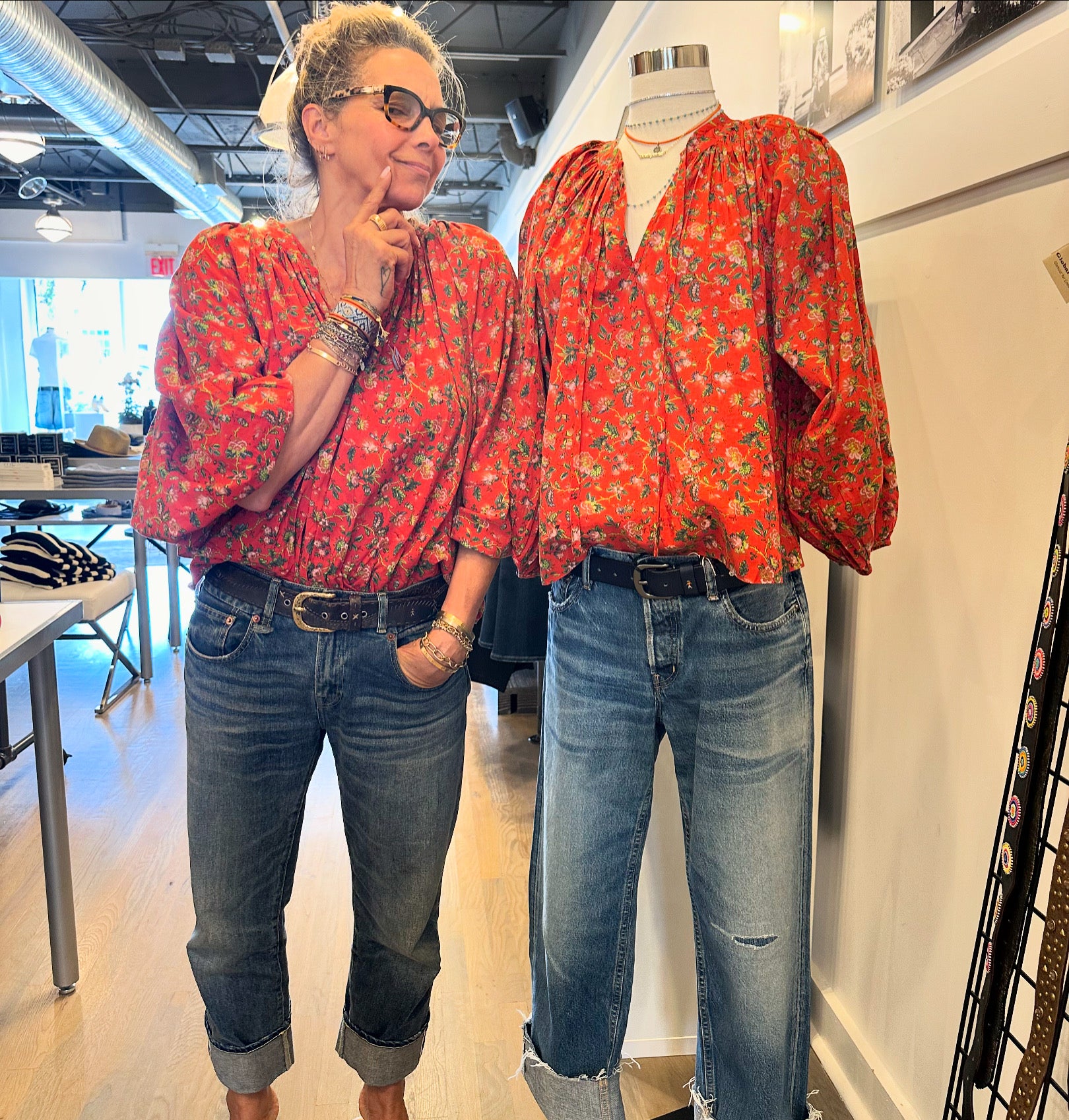 A person wearing a Rhode Red Flowered Poet Top made from organic cotton and blue jeans is posing next to a mannequin dressed in the same outfit inside a clothing store. The person has their hand on their chin, thoughtfully comparing their appearance with the mannequin.