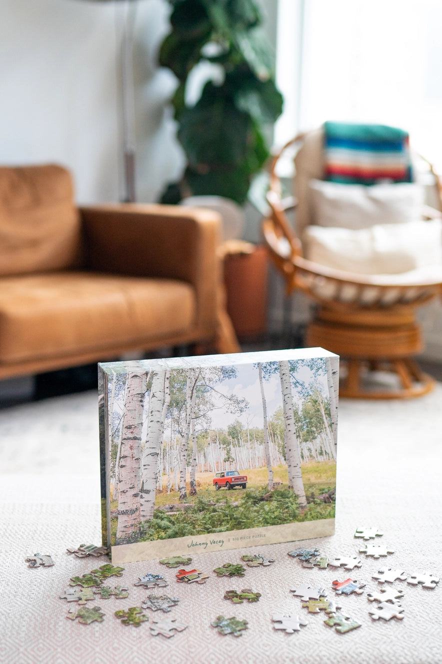 A completed Johnny Vacay 500 Piece Puzzle featuring a red car parked in a birch forest sits on the table. Scattered pieces of the puzzle are in the foreground, while in the background, there's a brown couch, a potted plant, and natural light streaming through the window—perfect for some family fun.