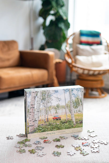 A completed Johnny Vacay 500 Piece Puzzle featuring a red car parked in a birch forest sits on the table. Scattered pieces of the puzzle are in the foreground, while in the background, there's a brown couch, a potted plant, and natural light streaming through the window—perfect for some family fun.