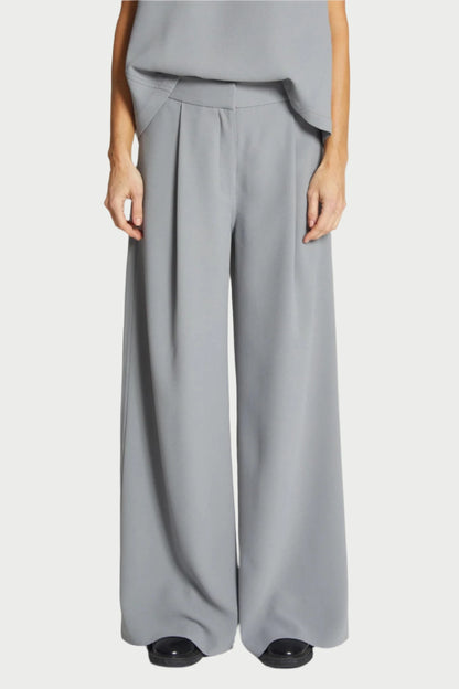 A person stands wearing the Saint Art NY Neve Crepe Pant in Powder Grey, featuring a wide-legged, loose fit. These versatile trousers have pleats at the waist and fall gracefully to the floor, covering their shoes. With relaxed tailoring, their hands rest casually at their sides as they pair the pants with a matching grey top.