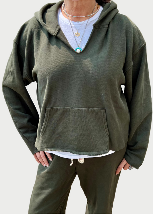 A person is wearing a comfy olive green hoodie and matching pants. The Les Tien Rachel Raw Crop Hoodie, featuring a front pocket, is layered over a white shirt. The individual accessorizes with layered necklaces while standing outdoors on a brick-paved area.